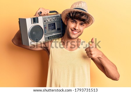 Young hispanic man holding boombox, listening to music smiling happy and positive, thumb up doing excellent and approval sign 