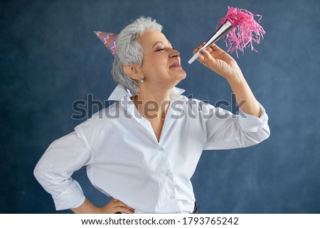 People, lifestyle, age and celebration concept. Isolated picture of happy overjoyed middle aged female wearing white shirt and pink cone hat blowing whistle while having fun at birthday party