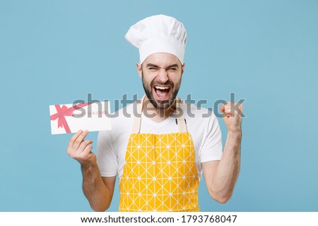Happy young bearded male chef or cook baker man in apron white t-shirt toque chefs hat isolated on blue background. Cooking food concept. Mock up copy space Hold gift certificate doing winner gesture