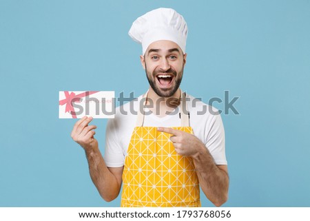 Excited young bearded male chef or cook baker man in apron white t-shirt toque chefs hat isolated on blue background. Cooking food concept. Mock up copy space. Point index finger on gift certificate