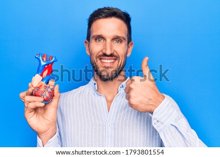 Young handsome man holding heart organ with veins and arteries over blule background smiling happy and positive, thumb up doing excellent and approval sign