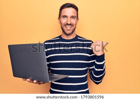 Young handsome man working using laptop standing over isolated yellow background pointing thumb up to the side smiling happy with open mouth