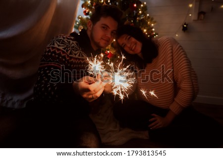 Young family with burning sparklers celebrating together in festive dark room. Happy New Year. Happy couple holding fireworks under christmas tree with lights.