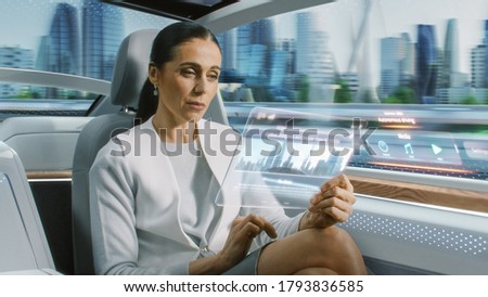 Attractive Senior Female Reading News on a Futuristic Transparent Tablet Computer with Augmented Reality Interface while Sitting on a Backseat of Autonomous Car. Self-Driving Van Rides on Public Road.