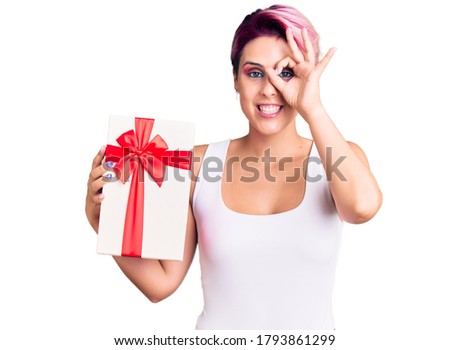 Young beautiful woman with pink hair holding gift smiling happy doing ok sign with hand on eye looking through fingers 