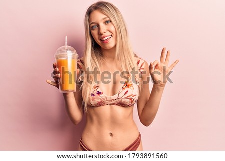 Young beautiful blonde woman wearing bikini drinking orange juice doing ok sign with fingers, smiling friendly gesturing excellent symbol 