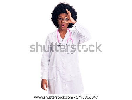 Young african american woman wearing doctor coat and stethoscope doing ok gesture shocked with surprised face, eye looking through fingers. unbelieving expression. 