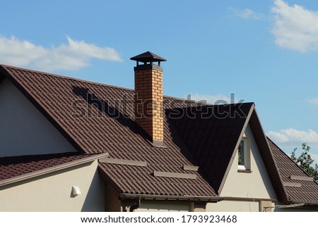 brown tiled roof of a gray private house with a window and one brick chimney against a blue sky