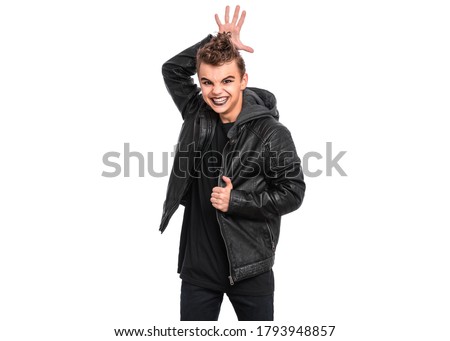 Rebellious teen boy dressed in black making horns with fingers on head, isolated on white background. Teenager in style of punk goth wearing leather jacket. Problems of transition age.