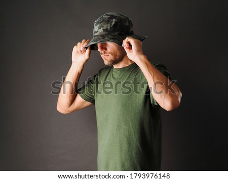 The man is wearing a green camouflage Panama hat and a green t-shirt on a grey background.