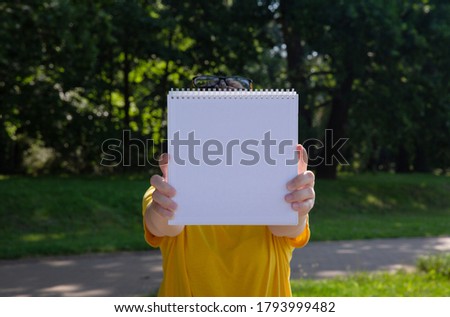  young woman in yellow t shirt holding a white book in front of her in the park on a sunny summer day. background is blurred. selective focus. High quality photo