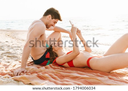 Happy young couple taking a selfie on the beach while laying on blanket