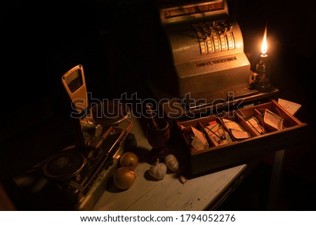 
cash register, scale, spices and old money lit by the flame of an old lamp on wooden table, "Low Key" image selective focus.
