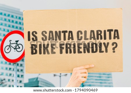 The question " Is San Clarita bike friendly? " on a banner in men's hand with blurred background. Transportation. Zero waste. Bicycle lane. Streets. City. Safety. Insecure. Road signs. Dangerous