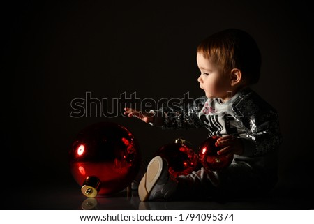 Toddler in gray boots and sparkling suit. She is playing with three red balls, christmas decoration, sitting on floor in twilight, against black background. New Year, holidays. Close up, copy space