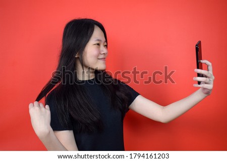 Portrait of Young beautiful asian woman using black T-shirt take a selfie picture using her smartphone with red isolated background