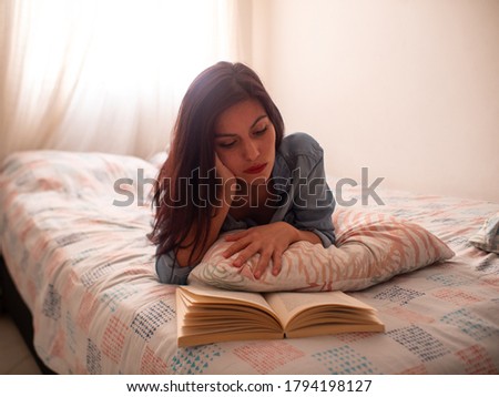 Young Hispanic Woman Reads a Book Lying on a Bed Supported by a Leaf Pattern Pillow