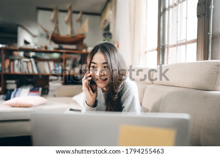 Young adult asian freelancer business woman working online via internet at home. Using mobile phone call with customer. Living room workspace background with window light.