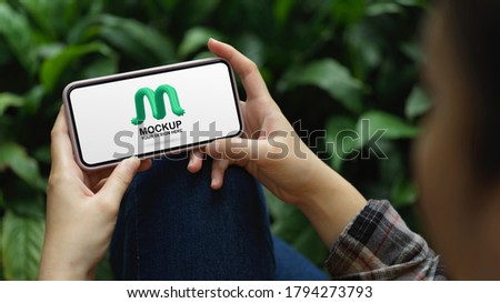 Close up view of a woman hands holding mock up horizontal screen smartphone while relaxed sitting in garden
