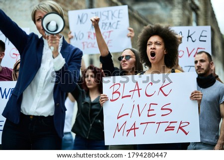 Large group of multi-ethnic people demonstrating against racism through city streets. Focus is on black woman shouting while holding a banner. 