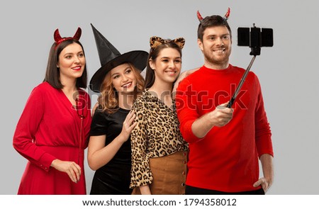 friendship, holiday and people concept - group of happy smiling friends in halloween costumes of witch, devil and cheetah taking picture by smartphone on selfie stick over grey background