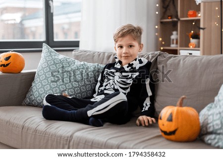 halloween, holiday and childhood concept - smiling little boy in party costume of skeleton sitting on sofa at home