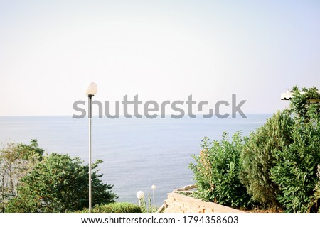 The sea in the background. Side green bushes. In the center of the photo
there is a lantern. The sky is pure color, blue. Photo color, horizontal. No people.