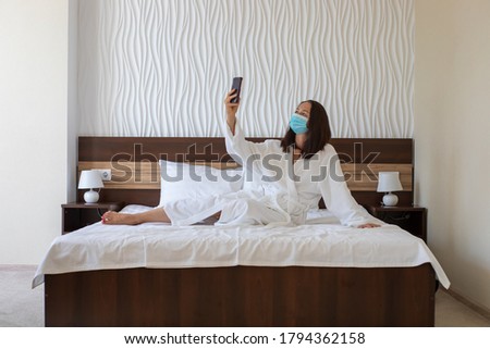 beautiful woman blogger wearing protective face mask with a smartphone lies in a large bed, takes a selfie photo