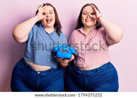 Young plus size twins holding vintage telephone smiling happy doing ok sign with hand on eye looking through fingers 
