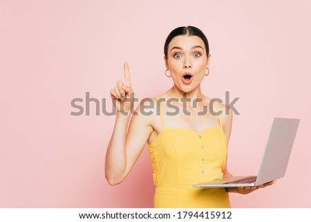 surprised brunette young woman using laptop and showing idea gesture on pink