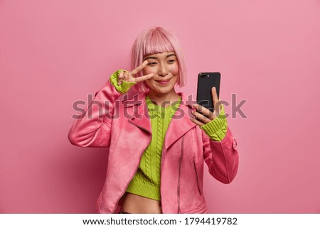Joyful Asian woman makes victory sign, takes selfie, holds modern smartphone, has pink hairstyle, looks in display of cellular, wears rosy jacket