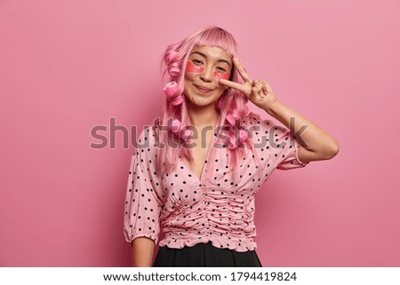 Pretty Asian woman wears hydrogel patches, hair curlers makes victory sign, dressed in blouse, pink background