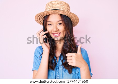 Young beautiful chinese girl wearing summer hat having conversation talking on the smartphone smiling happy and positive, thumb up doing excellent and approval sign 