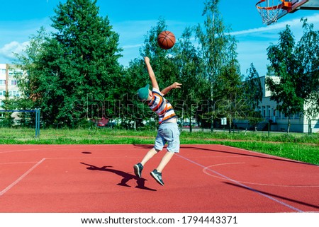 Attractive caucasian little basketball player jumps to throw the ball into the basket hoop on a outdoors red court