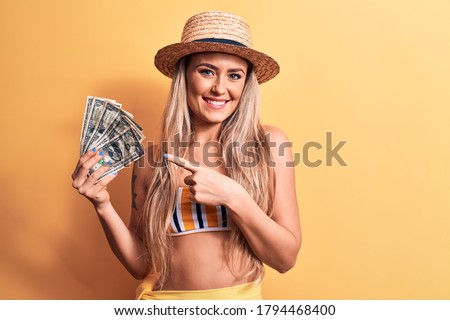 Young beautiful blonde woman wearing bikini and hat holding bunch of dollars banknotes smiling happy pointing with hand and finger