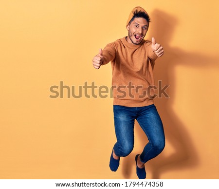 Young handsome latin man wearing casual clothes and wool cap smiling happy. Jumping with smile on face doing ok sign with thumbs up over isolated yellow background