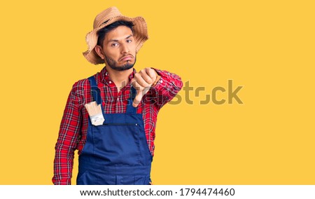 Handsome latin american young man weaing handyman uniform looking unhappy and angry showing rejection and negative with thumbs down gesture. bad expression. 