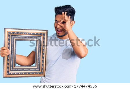 Handsome latin american young man holding empty frame smiling happy doing ok sign with hand on eye looking through fingers 