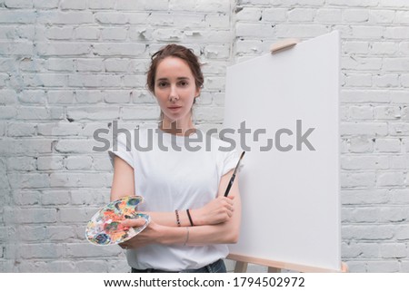 Beautiful woman posing, holding color palette with brush, artist in white T-shirt. White blank canvas, easel. Summer in city, brick wall background. Emotions comfort, pleasure, inspiration creativity.