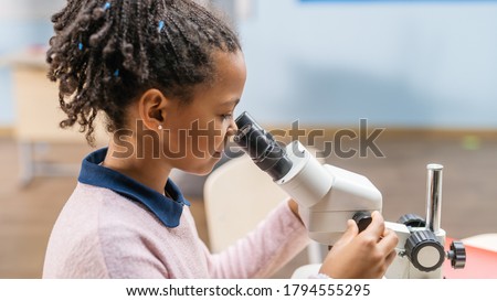 Portrait of Smart Little Schoolgirl Looking Under the Microscope. In Elementary School Classroom Cute Girl Uses Microscope. STEM (science, technology, engineering and mathematics) Education Program Royalty-Free Stock Photo #1794555295