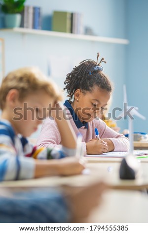In Elementary School Classroom Brilliant Black Girl Writes in Exercise Notebook, Taking Test and Writing Exam. Junior Classroom with Group of Bright Children Working Diligently and Learning New Stuff Royalty-Free Stock Photo #1794555385