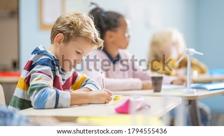In Elementary School Classroom Brilliant Caucasian Boy Writes in Exercise Notebook, Taking Test and Writing Exam. Junior Classroom with Group of Children Working Diligently and Learning New Stuff Royalty-Free Stock Photo #1794555436