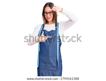 Beautiful brunette young woman dressmaker designer wearing atelier apron smiling making frame with hands and fingers with happy face. creativity and photography concept. 