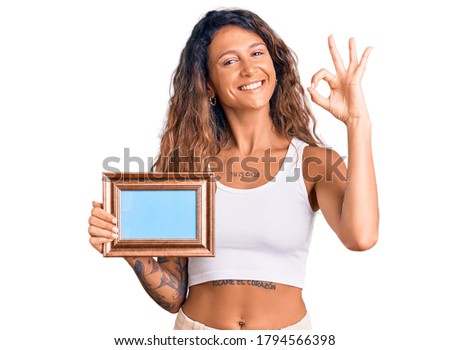 Young hispanic woman with tattoo holding empty frame doing ok sign with fingers, smiling friendly gesturing excellent symbol 