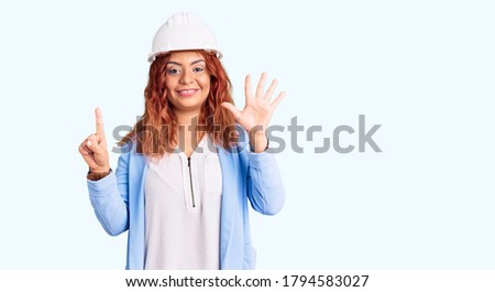 Young latin woman wearing architect hardhat showing and pointing up with fingers number six while smiling confident and happy. 