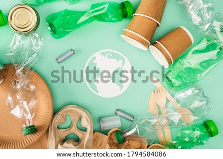 Top view of plastic bottles, accumulators and disposable tableware near paper earth sign on green background, ecology concept