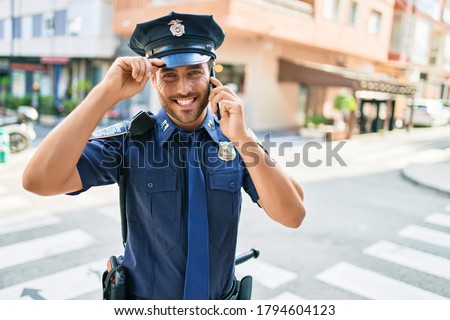 Young handsome hispanic policeman wearing police uniform smiling happy. Standing with smile on face having conversation talking on the smartphone at town street.