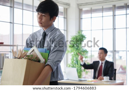 Asian young businessman fired from job standing sad and carrying his stuff box. The boss point to exit blur on background