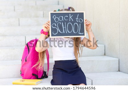 schoolgirl sitting, school backpack. He holds a sign in his hands with the inscription back that school and covers his face with it.