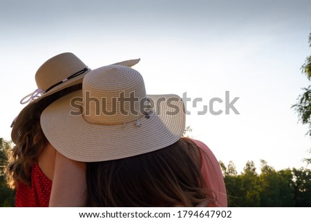Two girls with long hair and beige hats are embracing against the sky, their backs are turned.